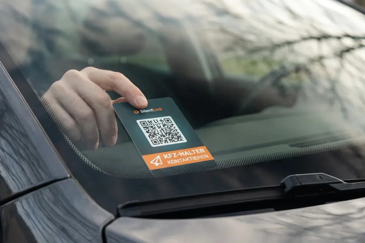 Eine Hand legt einen dunklen KFZ-Kontaktpunkt mit QR-Code und orangem Feld hinter die Windschutzscheibe