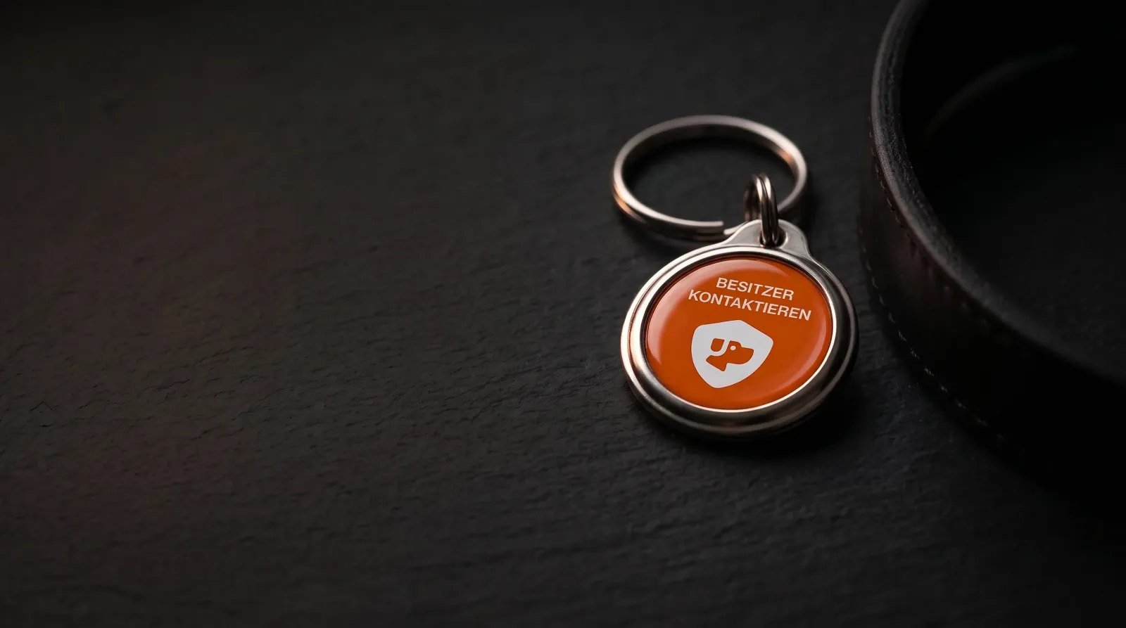 Die PetID liegt neben einem Lederhalsband auf dunklem Schiefer, weiches Streiflicht auf der orangefarbenen Front